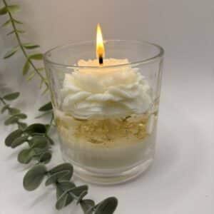 Kaars in doorzichtige glazen pot. Onderste laag is wit kaarsvet. Vervolgens doorzichtige gel met gouden schilfers in. Een witte pioenkaars als versiering bovenaan.