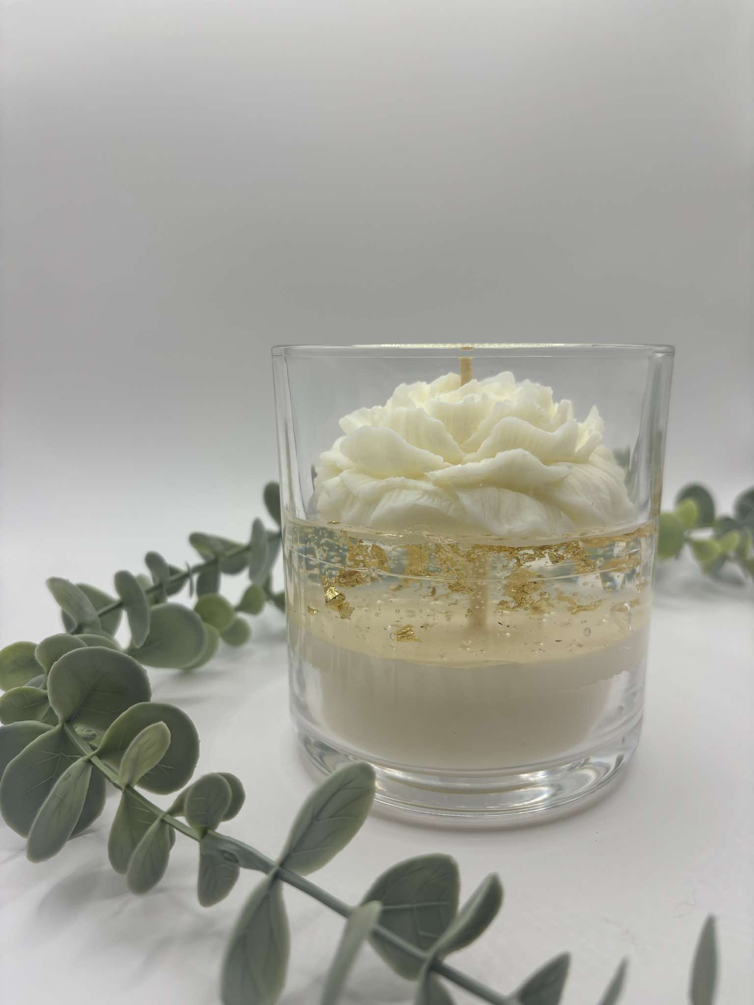 Kaars in doorzichtige glazen pot. Onderste laag is wit kaarsvet. Vervolgens doorzichtige gel met gouden schilfers in. Een witte pioenkaars als versiering bovenaan.